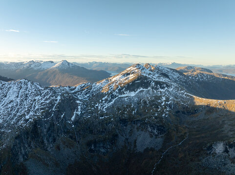Aerial view of the snow-capped peaks of Stetind mountain with rugged rocky ridges and distant fjords under a clear sky in Narvik Municipality, Nordland, Norway.