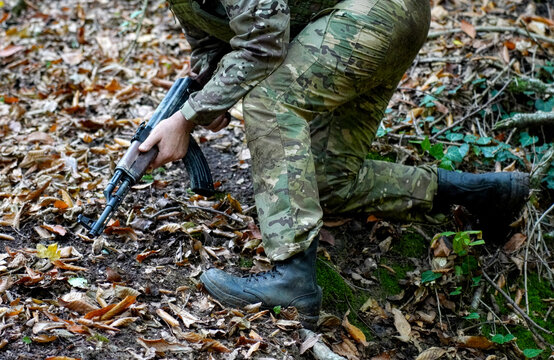Low angle shot of a soldier in camouflage gear kneeling on a forest floor covered in autumn leaves, holding an automatic weapon. Focus on tactical movement and military boots