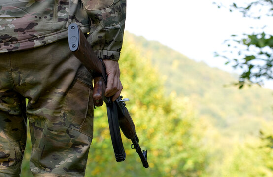 A close-up back view of a soldier or hunter wearing multi-terrain camouflage clothing, holding a wooden-stock assault rifle against a blurred green forest background