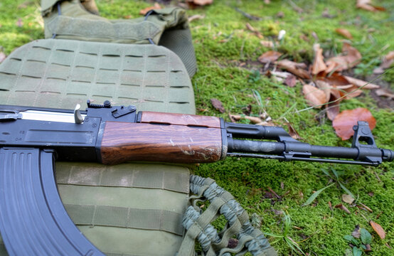 Assault Rifle Lying on Tactical Body Armor in Forest. Close-up of an automatic rifle with wooden stock resting on a green olive plate carrier (tactical vest) on a mossy forest floor