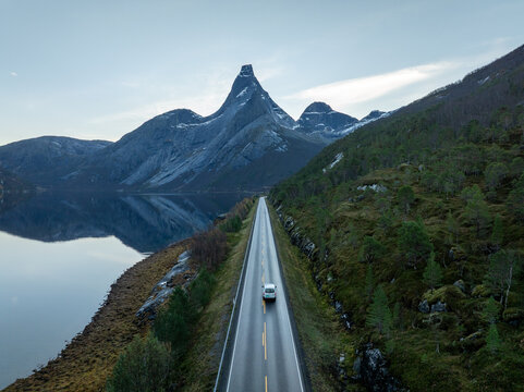 Aerial view of a white car driving on a straight road towards the iconic Stetind mountain peak reflected in the calm fjord waters in Narvik Municipality, Nordland, Norway.