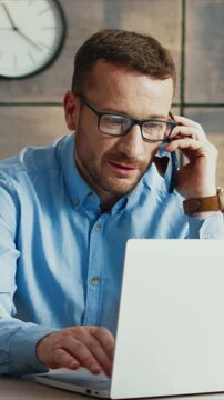 Caucasian man wearing glasses talking on smartphone while using laptop. Male checking information on screen and speaking with client. Working on task and focusing. Sitting at desk in light room.