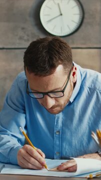 Vertical view of Caucasian man wearing glasses writing notes on paper at desk. Male holding pencil and focusing on task. Working on documents and concentrating. Sitting in room with clock behind.
