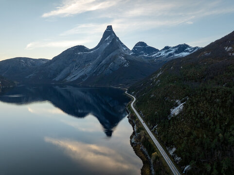 Aerial view of the iconic Stetind mountain peak reflecting in the calm waters of the fjord with a winding coastal road Narvik Municipality, Nordland, Norway.