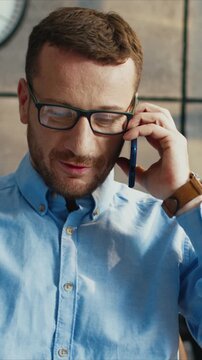 Caucasian man wearing glasses holding smartphone and speaking during call. Male looking down and focusing on conversation. Listening to client and thinking. Sitting in light room near desk.