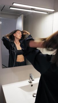 Attractive brunette woman wearing stylish black silk pajamas standing in modern bathroom, tying long hair into ponytail as looks at reflection in mirror
