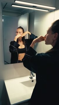 Woman in pajamas performing anti-aging facial massage exercises and facebuilding gymnastics, looking at her reflection in a modern bathroom mirror for skin care