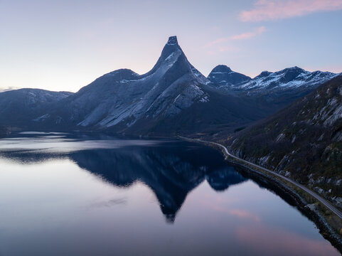 Aerial view of the iconic Stetind mountain peak reflecting in the calm waters of the fjord with snowy slopes and a coastal road at dawn in Narvik Municipality, Nordland, Norway.