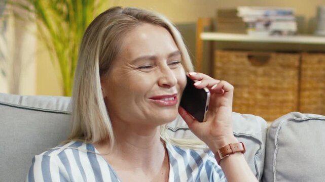 Caucasian woman smiling while talking on smartphone on sofa. Female holding phone near ear and reacting to call. Sharing good news and laughing. Spending time at home in cozy room.