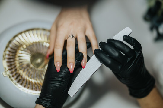 A manicurist files a client's pink ombr&eacute; nails while holding the hand steady. The salon technician carefully shapes the nails with a white emery board and black gloves for hygiene.