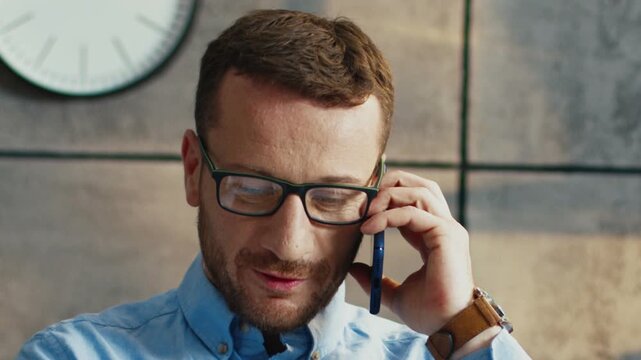 Close view of Caucasian man wearing glasses talking on smartphone. Male speaking with client. Holding phone near ear. Checking information while listening. Sitting in modern office room.