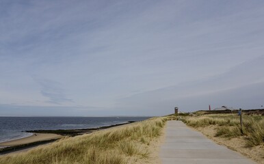 Fototapeta premium Wide-angle view of a coastal pedestrian path leading toward a lighthouse and observation tower on the sand dunes of Den Helder, Netherlands.