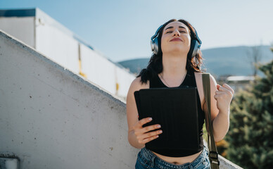 Young female student wearing headphones holds a tablet and smiles with her eyes closed. She enjoys music outdoors, raising a fist in a joyful, relaxed moment. © qunica.com