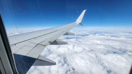 Fototapeta premium Airplane wing flying above white clouds against blue sky. View from aircraft window during flight. Travel and transportation concept for tourism and business trip.