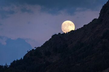 Fototapeta premium Full supermoon rising behind mountain slope, large bright moon over landscape in clear night sky