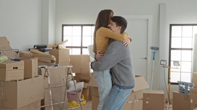 Young man lifts woman in tight hug among stacked moving boxes and open cartons in building entry room; new home joy.