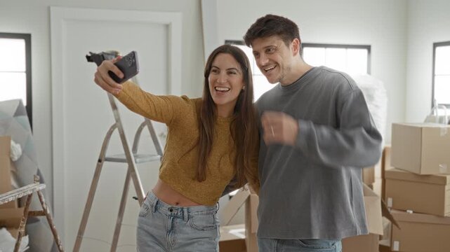 Young woman and man smile and hold smartphone for selfie amid packed boxes and ladder in a building; joy new home together.