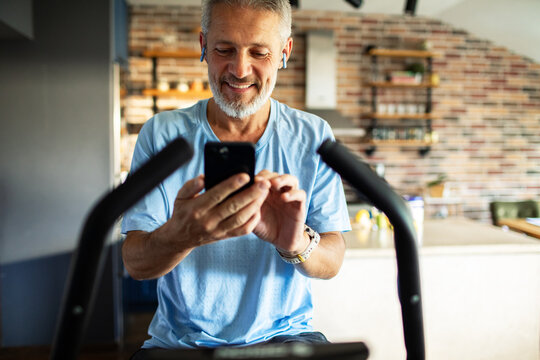 Smiling senior man using smartphone on exercise bike at home