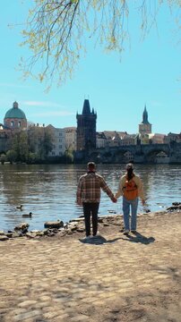 Couple walks hand in hand along the scenic Vltava River in Prague, enjoying the stunning architecture and vibrant atmosphere on a sunny day. Perfect for capturing travel memories.