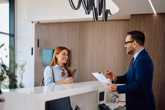 Corporate office receptionist assisting businesswoman with paperwork at check-in desk.