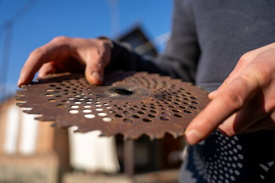 Person holding a rusty perforated metal cutting blade for brush cutter or trimmer, showing worn agricultural tool part outdoors.