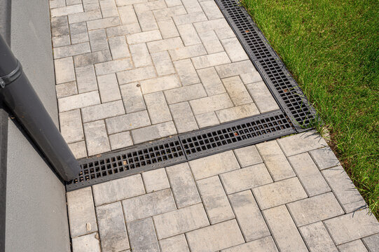 paved walkway with interlocking gray tiles, bordered by a black drainage grate. Adjacent to grass and a building corner, with a downpipe directing water into the drainage system