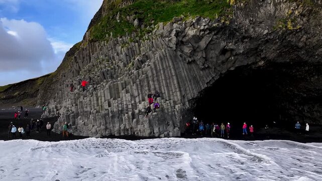 Aerial drone pulling away from Reynisdrangar basalt cliffs with tourists on black sand beach, Iceland