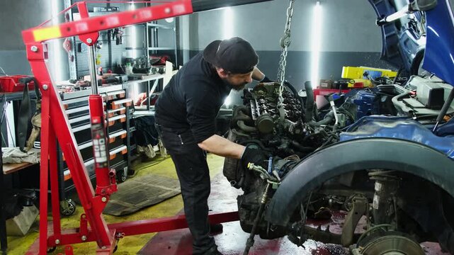 Young mechanic dismantles car engine with hydraulic hoist in repair shop slow motion. Man technician lifts motor with automated equipment in workshop