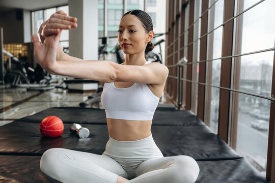 Asian female fitness enthusiast in white sportswear stretches arms while sitting on gym mat, with exercise equipment and large windows in the background