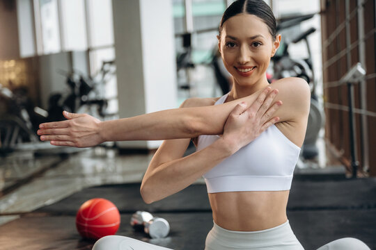 Asian female athlete stretches her arm while seated on a gym mat, with fitness equipment and a basketball visible in the background