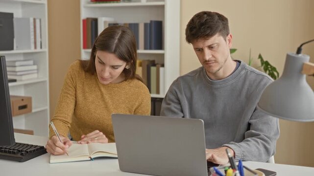 Young man and woman working on laptop, woman writing in notebook with pen while man types and smiles toward camera in building workspace; friendly collaboration focus.