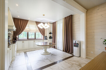 Fototapeta premium Elegant kitchen-dining area with marble flooring, white cabinetry, round dining table, chandelier, large windows with brown curtains, light wood walls, and water dispenser, bright and cozy.