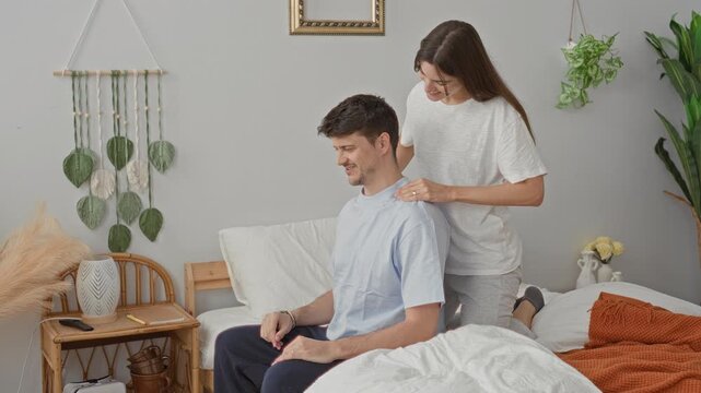 Young couple smiling, woman massaging man's shoulder on bed in cozy bedroom with blanket and pillow; intimacy comfort tenderness.