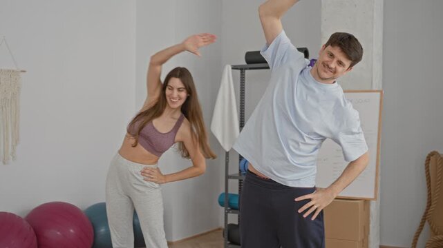 Woman stretching with arms raised in a side bend, smiling next to exercise balls, yoga props, whiteboard and shelving inside a home studio; together joy.