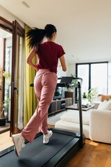 Fototapeta premium Young woman running on treadmill near glass doors at home