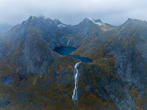 Aerial view of Lofoten mountains with a dark blue alpine lake and a waterfall cascading down rocky slopes under a cloudy sky Nordland, Norway.