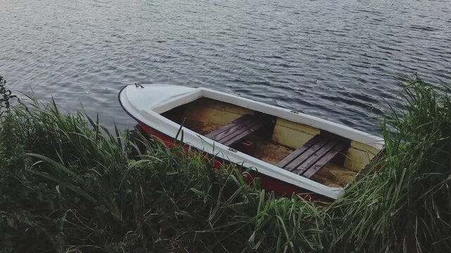 Old wooden rowboat moored by the grassy shore, gently bobbing on the rippling surface of a lake. A calm and lonely natural scene