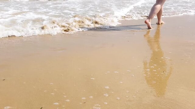 Woman's bare feet and legs walking along the wet sand of a beach. The gentle sea waves wash over her feet as she walks on the shoreline in Pineta, Lignano Sabbiadoro, Italy