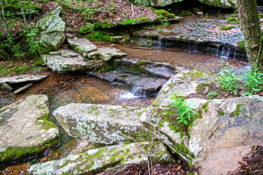 Water flows gently cascading along the river rolling over rocks and ledges. Boulders and green foliage surround the waters. Bokeh.