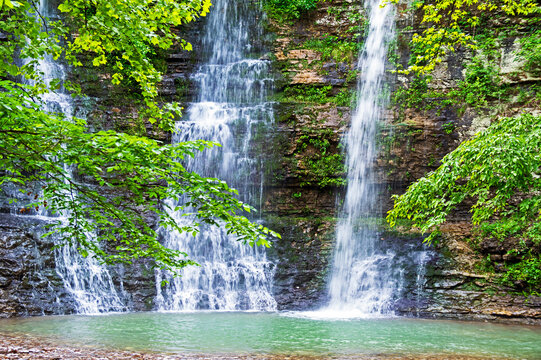 A slow shutter speed and a triple waterfall reveals a delightful scene in nature with a cliff wall and green foliage surrounding a pool of fresh water in Arkansas. Bokeh.
