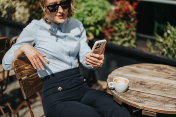 Fototapeta premium A woman in business attire sits at a wooden cafe table holding a smartphone. She relaxes with a latte while checking her phone in bright sunlight.