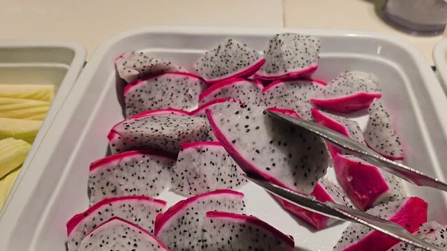 Fresh sliced dragon fruit in white tray with metal tongs at buffet, close up of pitaya pieces with black seeds and pink skin in self service fruit bar display