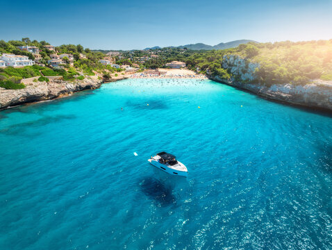 Aerial view of boat and serene sandy beach cove in Manacor, Balearic Islands. Mallorca, Spain. A solitary yacht floats on crystal-clear turquoise waters, surrounded by lush greenery and coastal villas