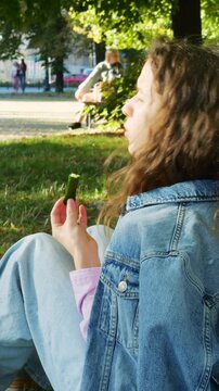 Attractive teenage girl eats fresh cucumber and chats cheerfully with friends. Child glances around at people strolling through city park. Vertical video