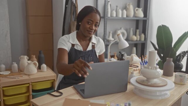 Black woman potter smiling and reaching for laptop lid while seated among clay pots, brushes and tools in a studio; creativity serenity.