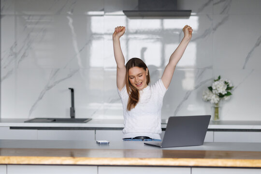 Young woman at a kitchen table expressing excitement with her arms raised and laptop open, working online