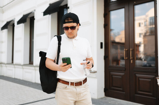Man Checking Phone Holding Water Bottle On Street, Wearing Sunglasses And Backpack, Focused On Notifications, Modern