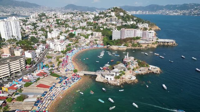 Stunning aerial drone footage captures the lively Caleta and Caletilla beaches in Acapulco, Mexico. Sunny day with tourists enjoying the bay, boats, and coastal cityscape.