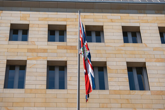 British Union Jack flag hanging on a flagpole in front of a modern sandstone government building facade under clear blue sky.