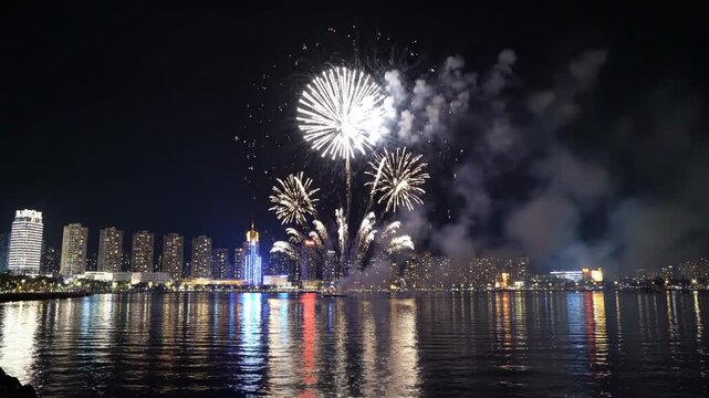 Bright white fireworks exploding over a city skyline at night with reflections on the water.